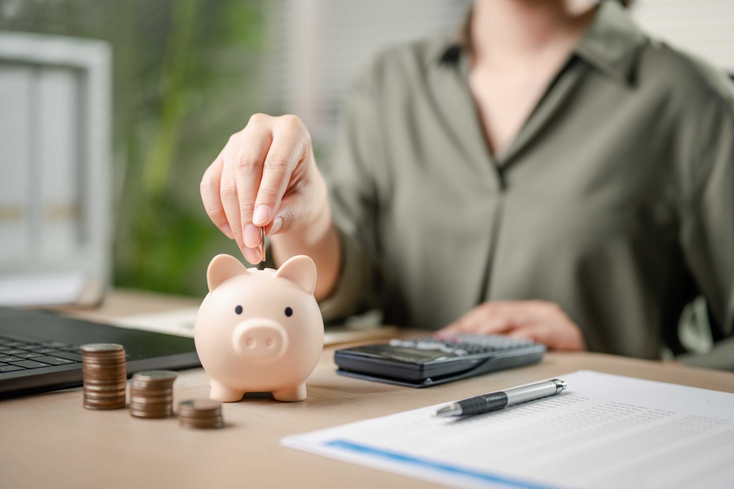 Person putting coins into a pink piggy bank that sits on a desk.